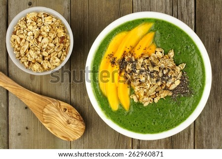 Green smoothie bowl with mangoes, granola, almonds and chia seeds, overhead view on wood with spoon