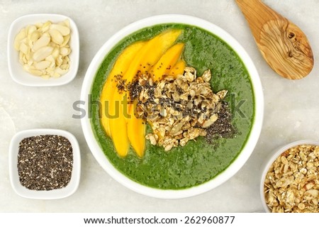 Green smoothie bowl with mangoes, granola, almonds and chia seeds, overhead view on white granite
