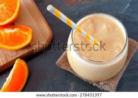 Healthy orange smoothie in a glass with striped straw and fresh fruit slices, downward view on slate