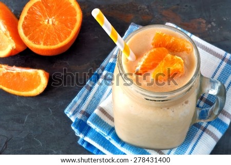 Orange fruit smoothie in a mason jar glass resting on checked cloth with fresh orange slices over a slate background