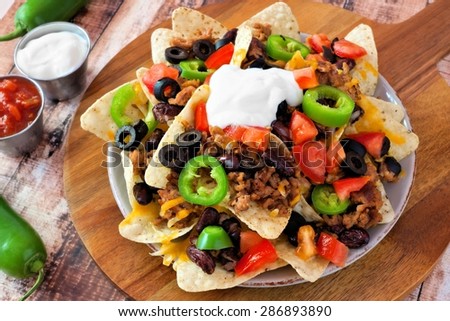 Mexican nacho chips topped with sour cream, ground meat, jalapenos, tomatoes, beans and melted cheese on a wooden paddle board