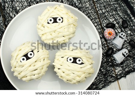 Group of Halloween mummy cupcakes on white plate with candies on black cloth background