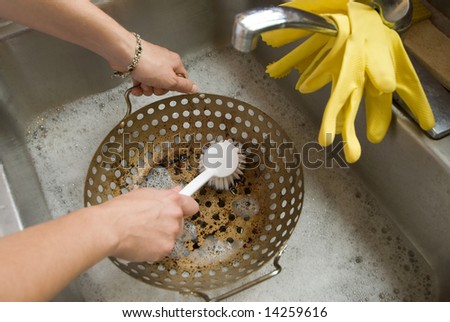 a woman washes a colander in a stainless steel sink by scrubbing it with a white plastic brush