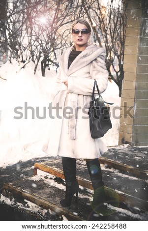 Glamorous woman in sunglasses standing on the porch in the winter in a white fur coat