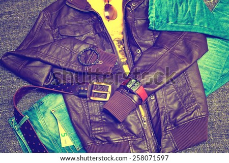 clothing items and accessories: blue jeans with a leather belt, leather jacket, T-shirt, watches, sunglasses and a bracelet on a hand