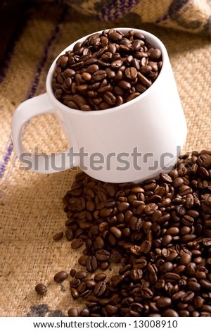 A white cup full of roasted coffee beans on a burlap coffee bean bag.  Background with copy space