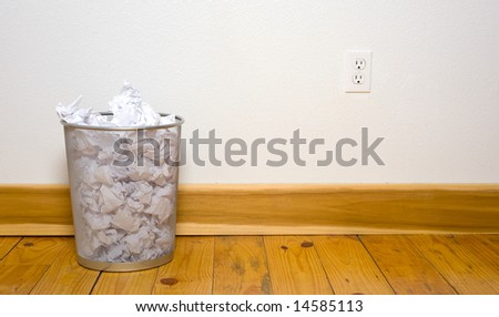 a wire mesh office trash can with crumpled paper on a wooden floor with a white wall, includes copy space