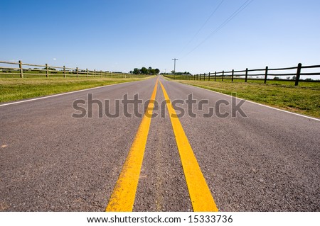 A long road bordered by farm fences and telephone wire against a blue sky