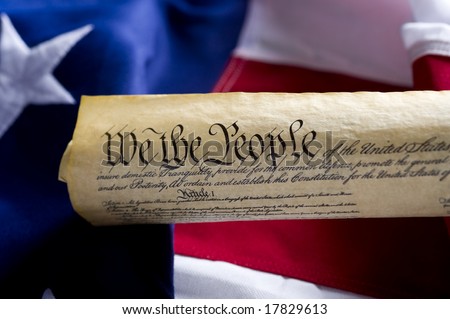 A copy of the United States of America Constitution scroll in front of a large American Flag, focus is on the word people.