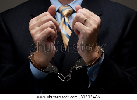 A man in a blue business suit holding hands in handcuffs in front of his chest- white collar or corporate crime