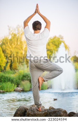 Young man practicing yoga, doing tree pose and turning back upon cmera. With a beautiful green scenery of park on background