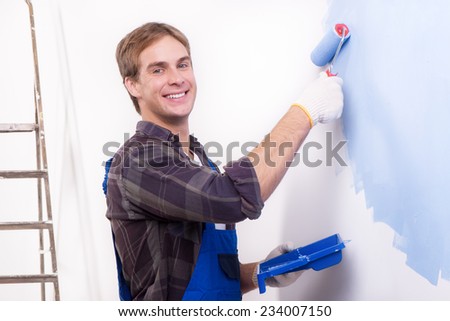Young smiling repairman painting wall by roller and looking at the camera