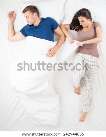 Young couple sleeping in big white bed. Top view photo. Man and woman can not divide a blanket. Angry woman trying to take a blanket