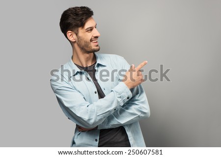 Handsome young man smiling and pointing at something with finger, standing on grey background