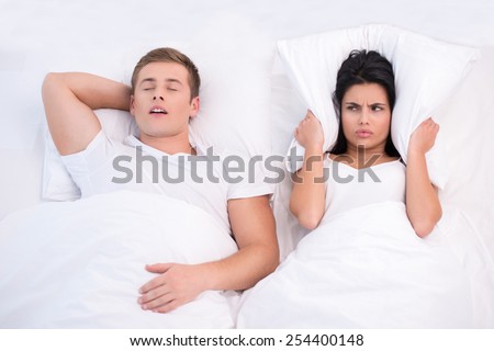 Top view photo of young couple lying in bed under white blanket. Woman is angry because of snoring husband