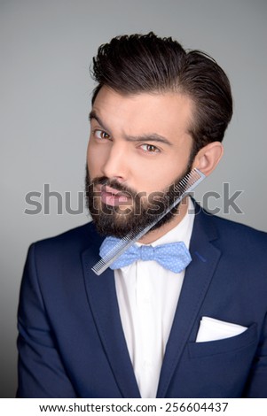 Photo of handsome stylish man with beard and comb in it. Man wearing classic dark blue suit and a blue bow tie. Man looking at camera