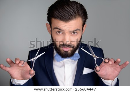 Photo of handsome stylish man with beard. Man wearing classic dark blue suit and a blue bow tie. Man holding scissors and provocatively looking at camera