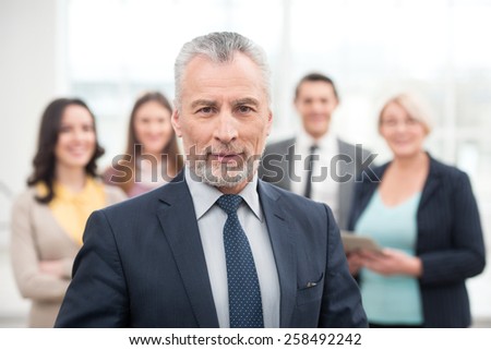 Businessman looking at camera. His team standing behind. Office interior with big window