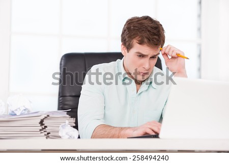 Young sad businessman looking at papers and folders while sitting at his black leather office chair. Office interior with window