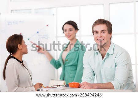 Young smiling businessman looking at camera. His creative team working with diagram on background. Office interior with window