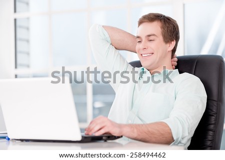 Young smiling relaxed businessman working with laptop and sitting at his black leather office chair. Office interior with window