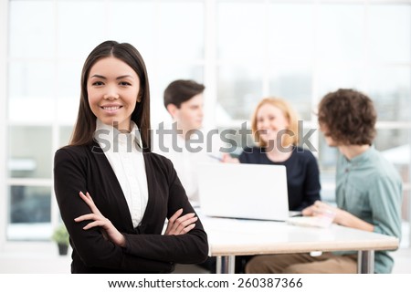 Businesswoman looking at camera with her business team on background. Office interior with window. Concept for teamwork