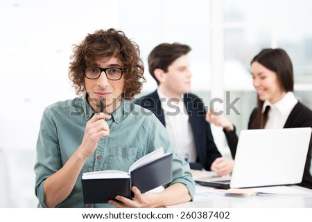 Young casual businessman holding notebook with his business team on background. Office interior with window. Concept for teamwork