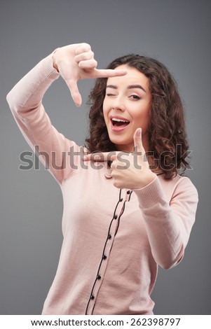 Beautiful young woman winking and making hand frame, standing on grey background