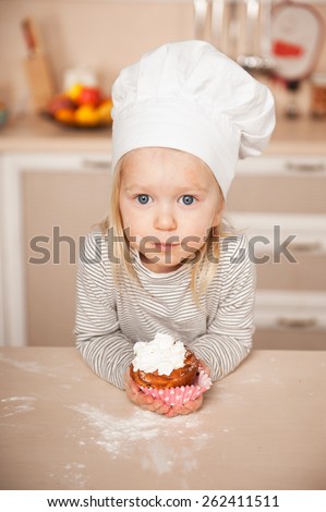 Little cute girl with chef hat holding cake and looking at camera. Kitchen interior. Concept for young kitchen hands