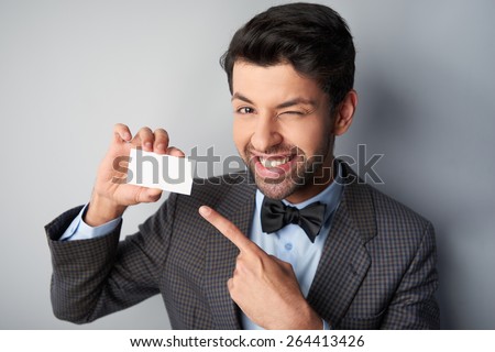 Portrait of positive casual young man wearing jacket and bow tie. Man smiling and pointing at blank visiting card