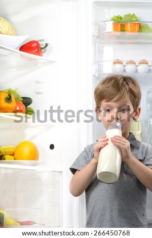 Little cute boy drinking milk near open fridge. Vegetables and fruits in the refrigerator