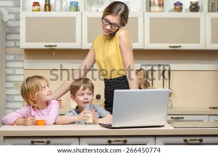 Young business woman early in the morning with her little children. She using laptop and phone. Kitchen interior. Concept for busy mother
