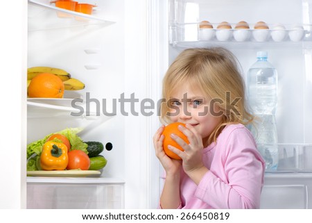 Little cute girl looking at camera and holding orange near open fridge. Vegetables and fruits in the refrigerator
