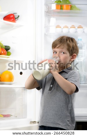 Little cute boy drinking milk near open fridge. Vegetables and fruits in the refrigerator