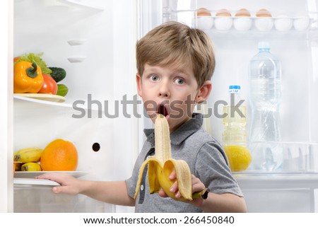 Little cute boy looking at camera and eating banana near open fridge. Vegetables and fruits in the refrigerator