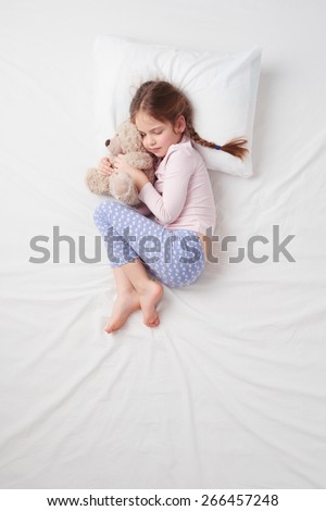 Top view photo of little cute girl sleeping on white bed with teddy bear. Quiet Foetus pose. Concept of sleeping poses