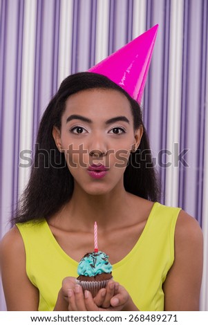 Photo of young african-american woman looking at camera. Woman wearing birthday hat and holding small cupcake with candle. Concept for happy birthday