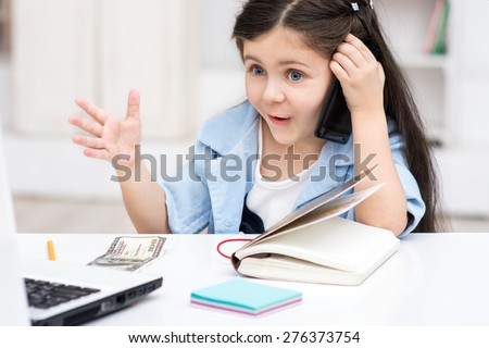 Funny picture of little dark-haired girl playing role of business woman. Girl sitting at table and amazedly using phone. Office interior as a background