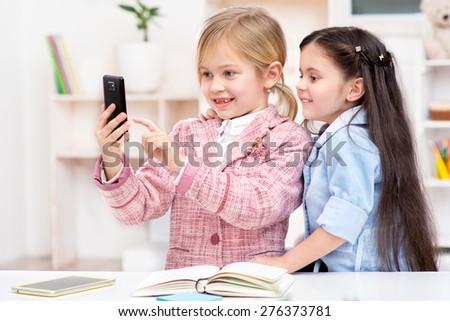 Funny picture of two little cute girls playing role of business women. Girls smiling and making photo by phone. Office interior as a background