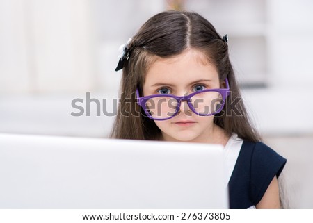 Funny picture of little dark-haired girl playing role of business woman. Girl wearing glasses. Girl sitting at table and looking at camera from behind laptop. Office interior as a background