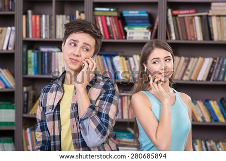 Handsome boy and beautiful girl smiling and using phones. They sitting in library with a lot of books. Concept for teenagers