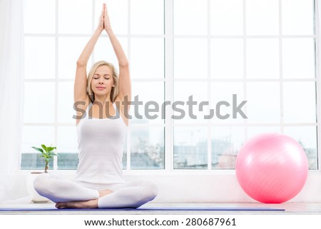 Nice photo of beautiful woman practicing yoga. Woman meditating with her eyes closed while doing lotus pose. White interior with large window and pink fitness ball