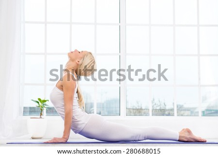 Nice photo of beautiful woman practicing yoga. Woman meditating with her eyes closed while doing cobra pose. White interior with large window