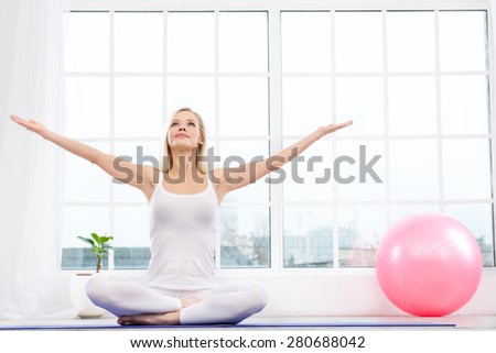 Nice photo of beautiful woman practicing yoga. Woman meditating with her hands up while doing lotus pose. White interior with large window and pink fitness ball