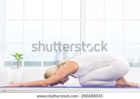 Nice photo of beautiful woman practicing yoga. Woman meditating with her eyes closed while doing child\'s pose. White interior with large window