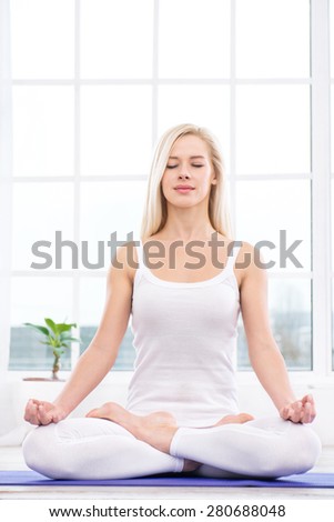 Nice photo of beautiful woman practicing yoga. Woman meditating with her eyes closed while doing lotus pose. White interior with large window