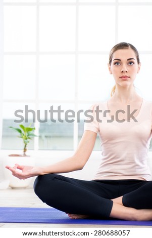 Nice photo of young woman practicing yoga. Woman meditating while doing lotus pose. White interior with large window