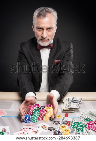 Man in years sitting at poker table, making bets and looking at camera. The chips and money are on table