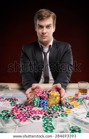 Pleased young man sitting at poker table, making bets and looking at camera. The chips, cards and money are on table