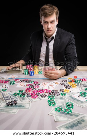 Young man sitting at poker table, nervously holding his hand on money and suspiciously looking aside. The chips, cards and money are on table